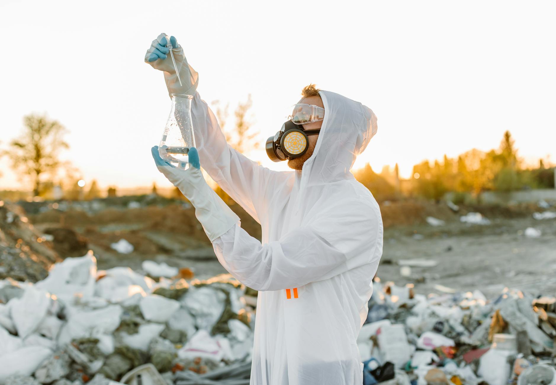 Scientist in protective gear collecting samples at a waste site during sunset.