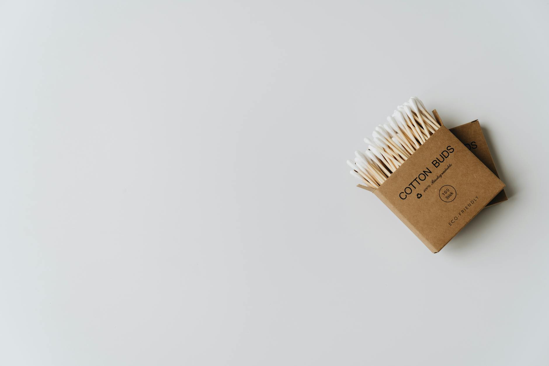 Minimalist image of eco-friendly cotton buds in cardboard packaging on a white surface.