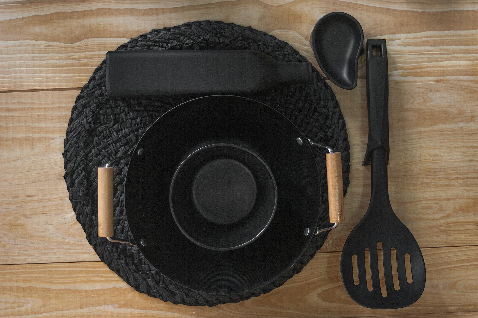 Black ceramic bowl, bottle, and utensils arranged on woven mat and wooden surface from above.
